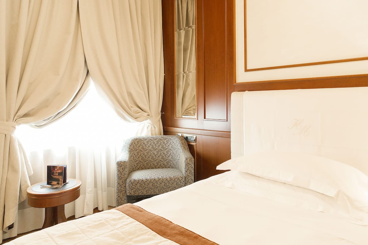 Una habitación de hotel con cama, mesita de noche con un libro, una silla y ventana con cortinas.
