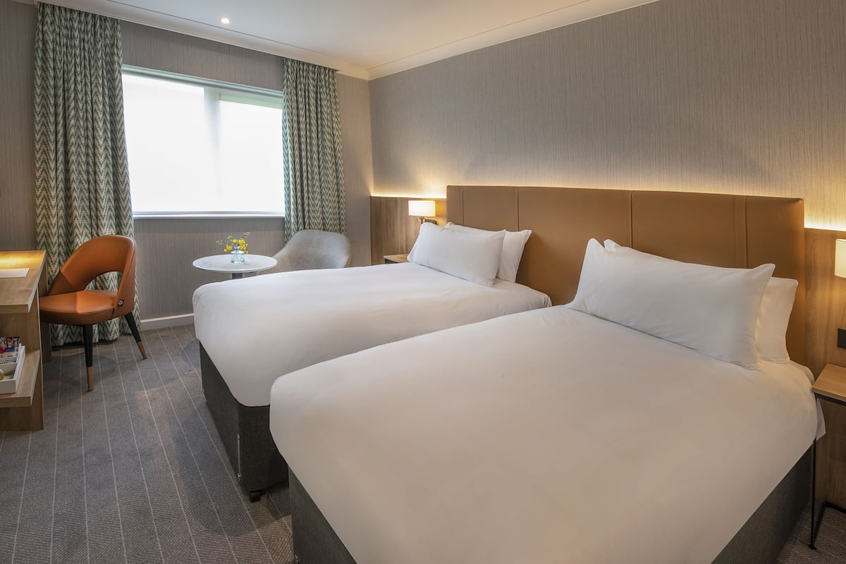 A hotel room with two beds, a small table, a chair, and a window with curtains.