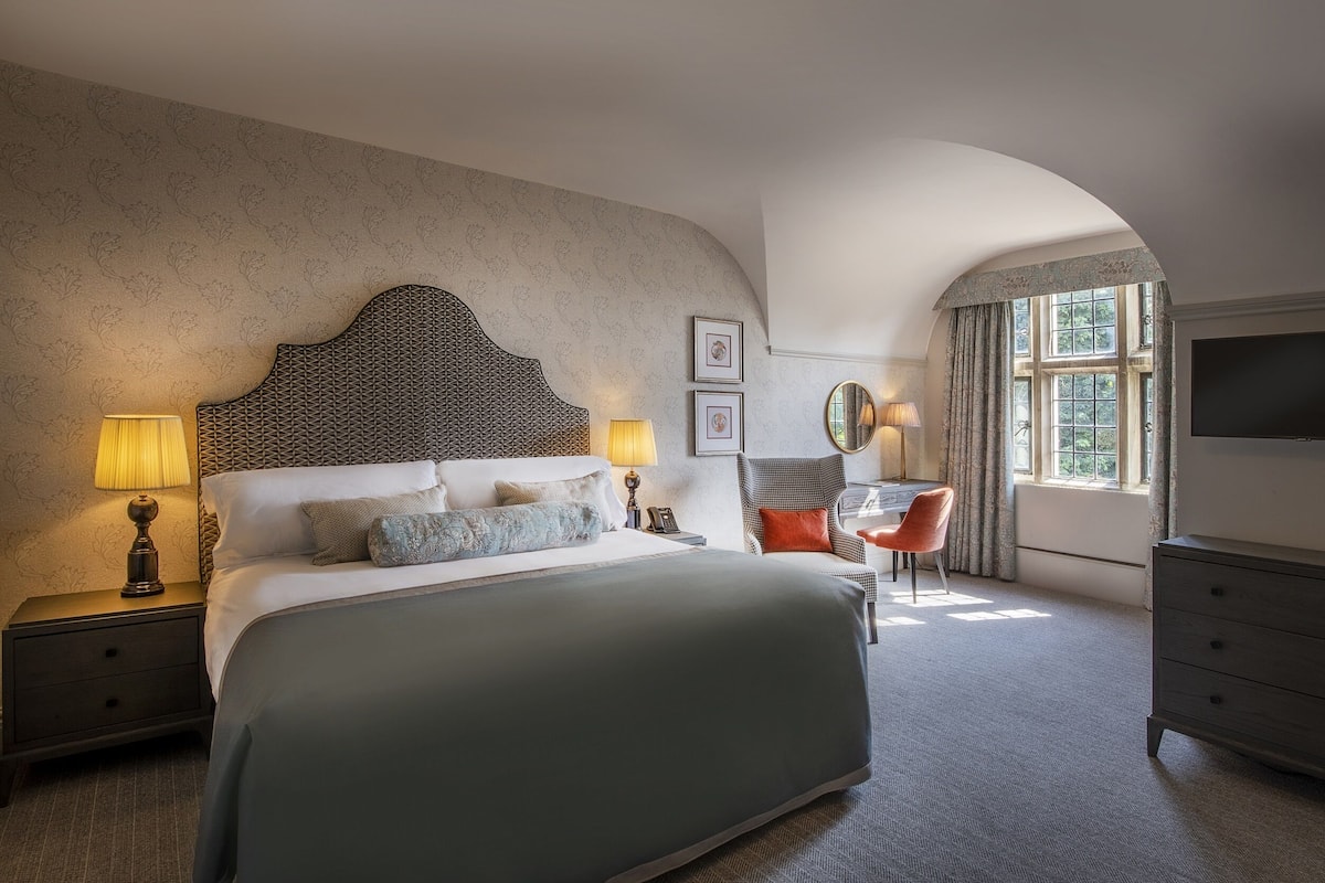 A hotel room with a large bed, two bedside lamps, a chair, a TV, and a window with curtains.