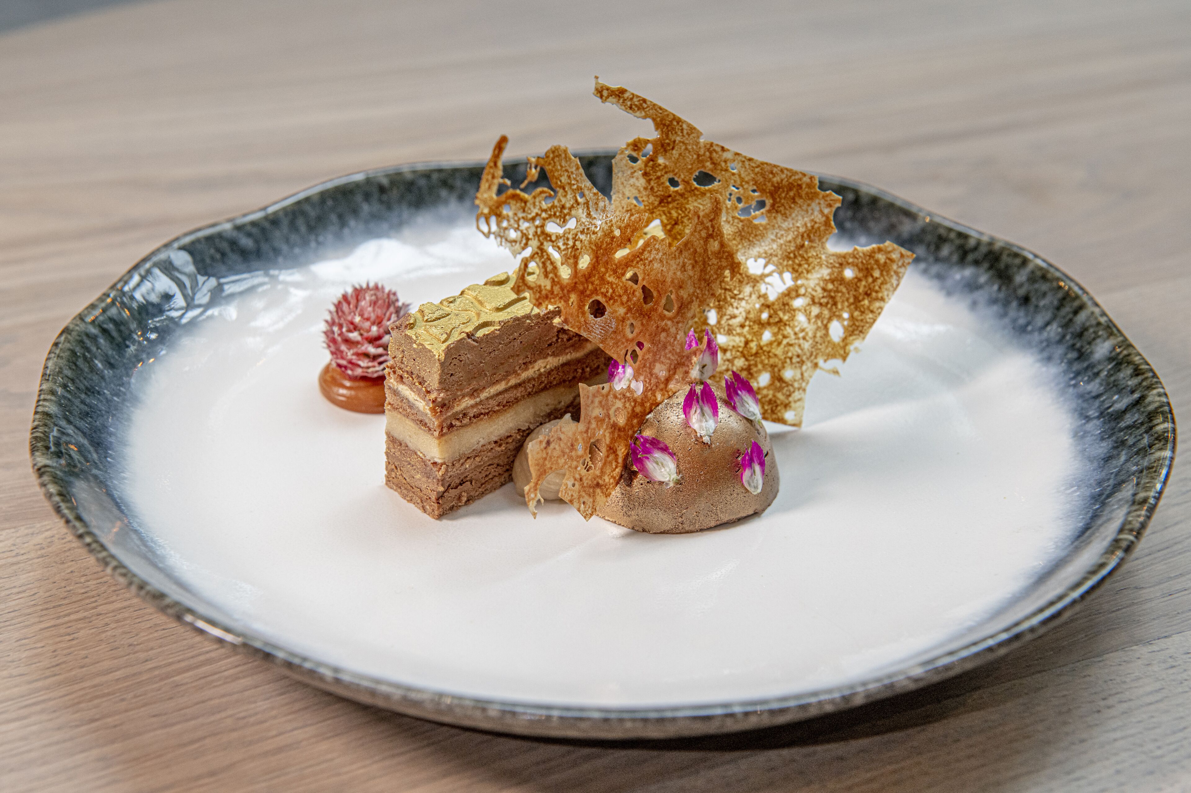 Une assiette à dessert comprenant une part de gâteau à plusieurs couches, une pâtisserie en forme de sphère et des éléments décoratifs, le tout présenté sur une assiette en céramique.