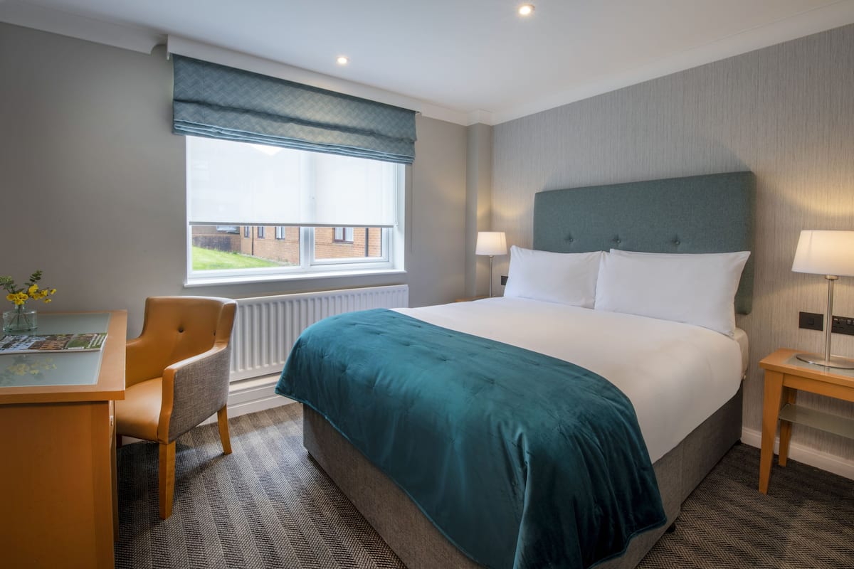 A hotel room with a bed, a desk, a chair, a window with a view, and a lamp.
