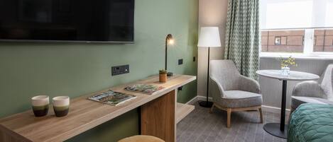 Habitación de hotel con televisión, escritorio con dos tazas, lámpara, dos sillas, una mesa redonda y una cama.