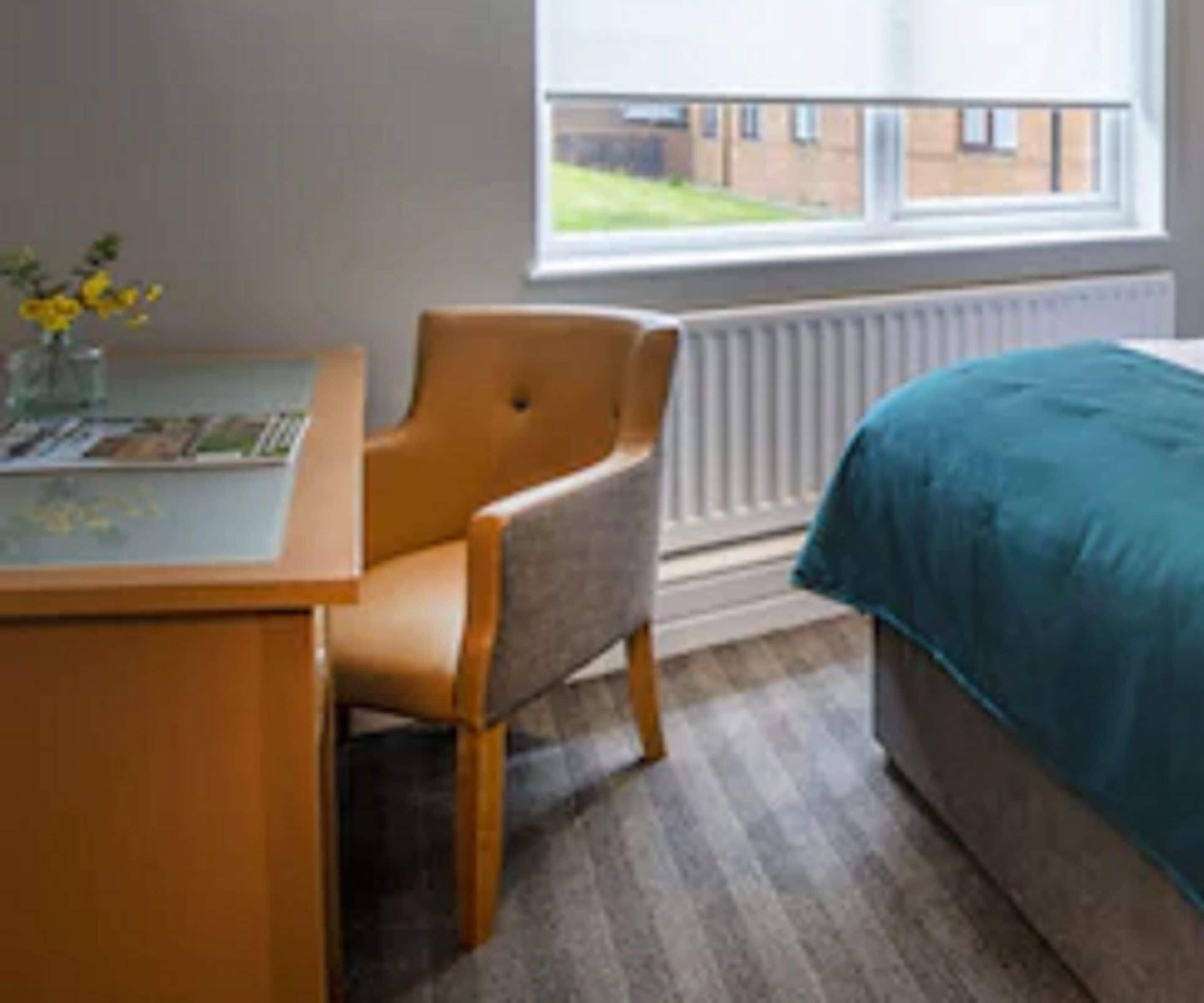 Ein Zimmer mit einem Bett, einem Sessel, einem Schreibtisch mit Blumenvase und einem Fenster mit Blick auf Gebäude.