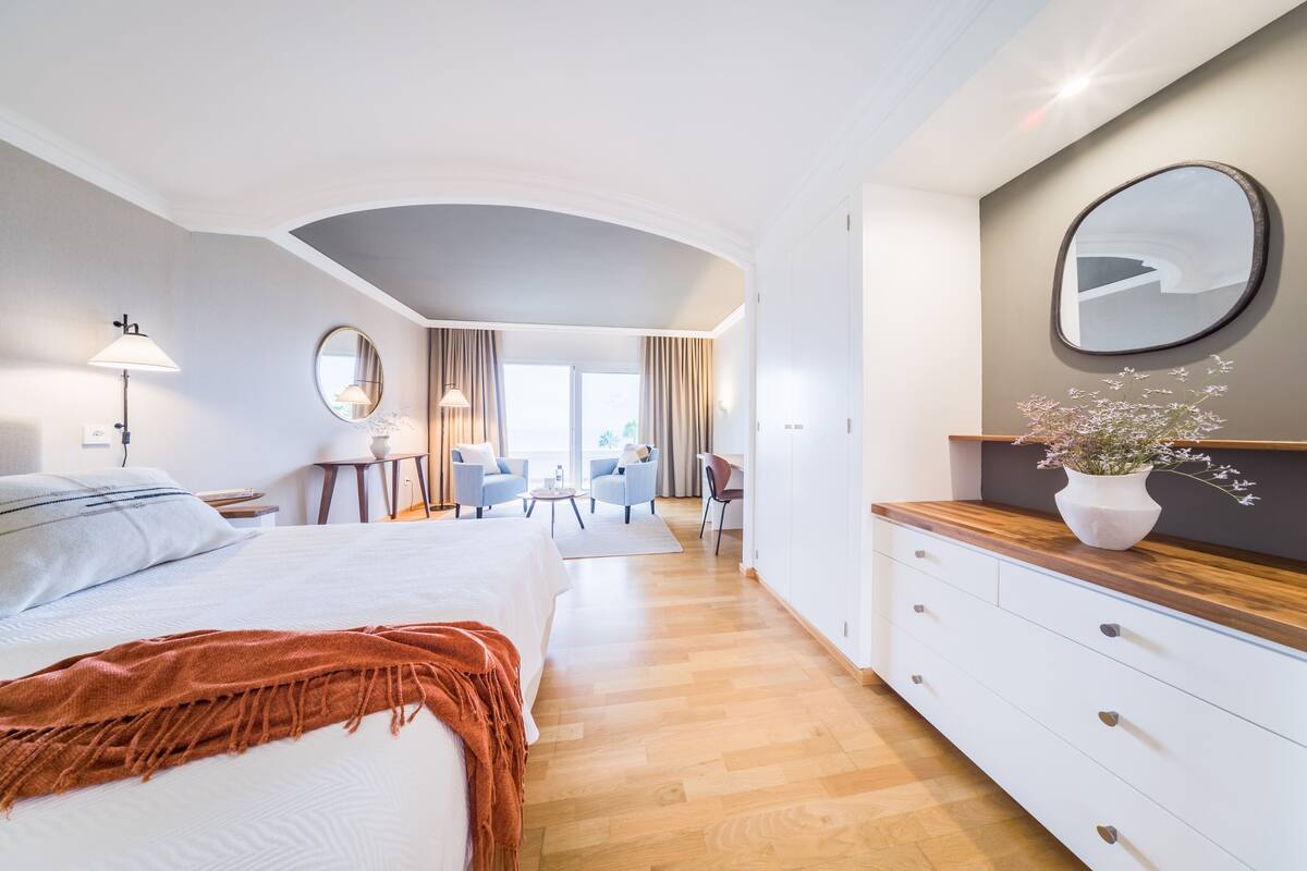 Ein modernes Hotelzimmer mit einem großen Bett, einem Schreibtisch, zwei Stühlen, einem Spiegel und einer Holzkommode.