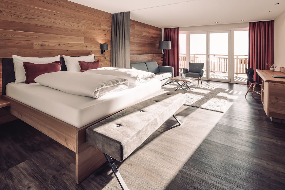 A modern hotel room with a wooden headboard, a large bed with white linens, a bench at the foot of the bed, a sofa, a desk, and a balcony with red curtains.