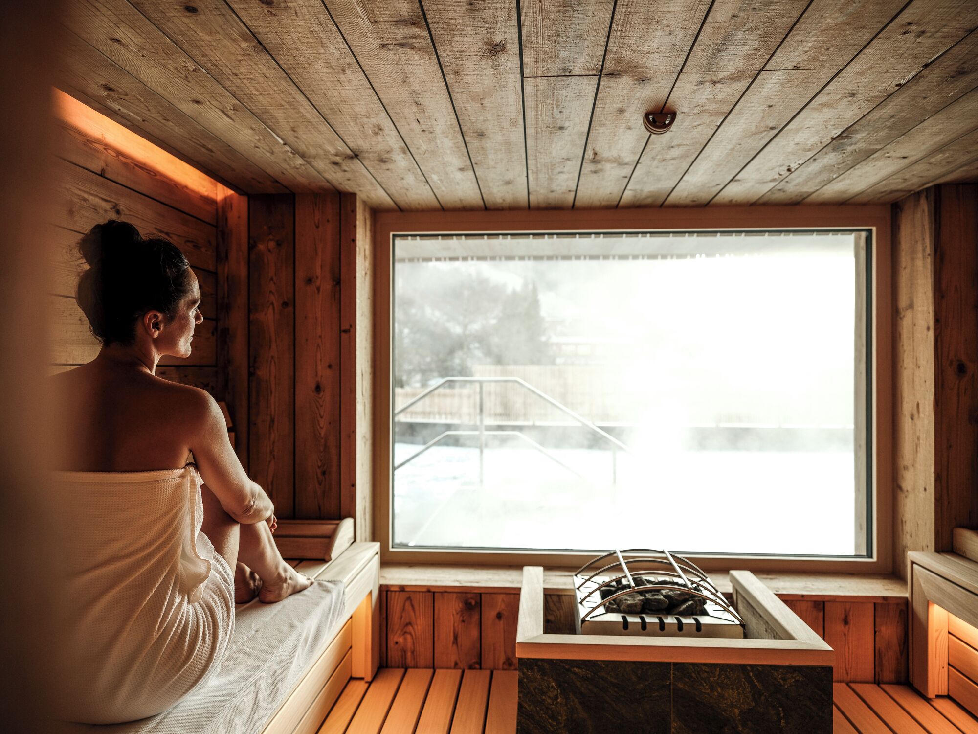 A wooden cabin with a sauna, a window with a view of a wooden deck, and a stone bench.