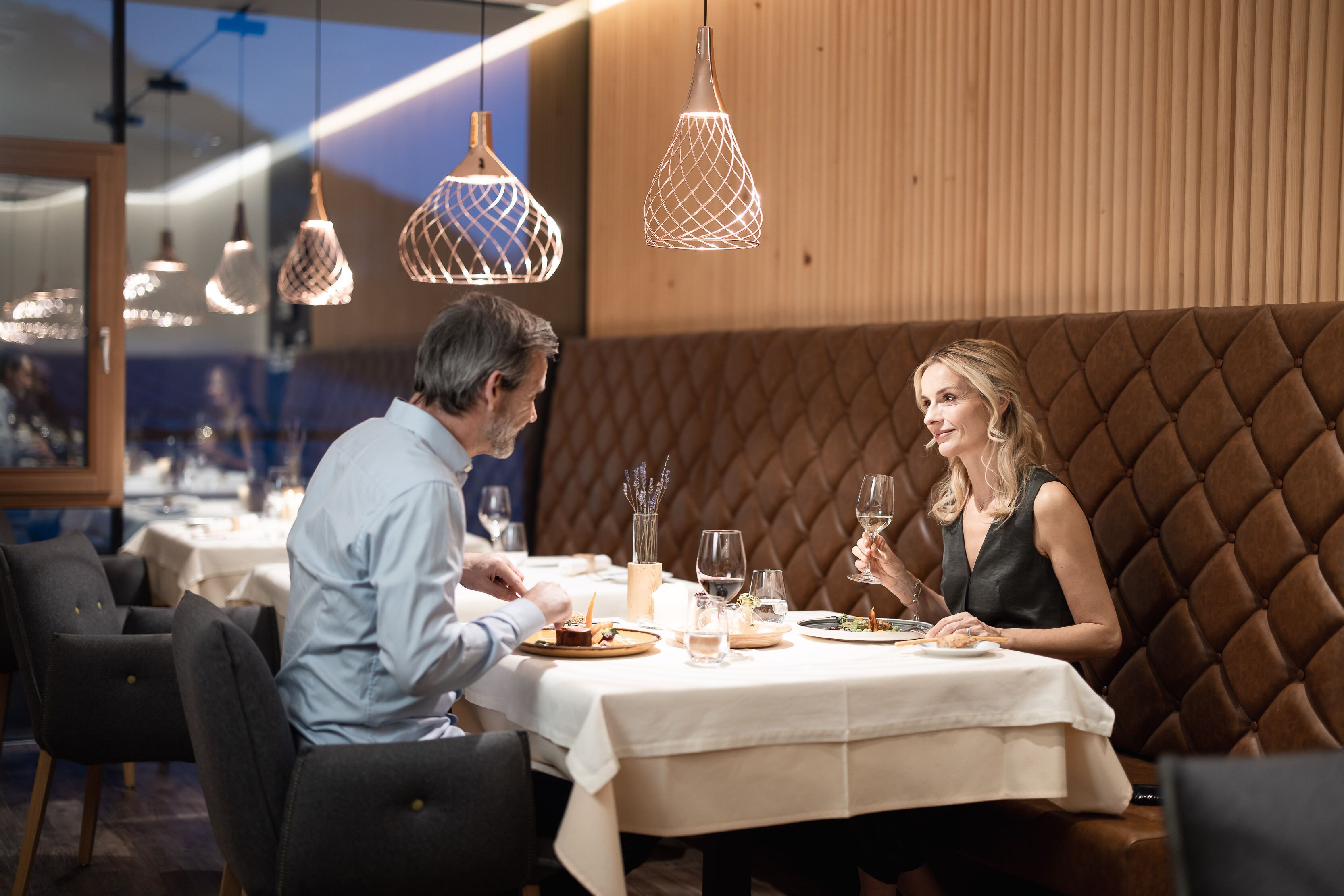 A couple dining at a restaurant with a tufted booth, pendant lighting, and a white tablecloth.