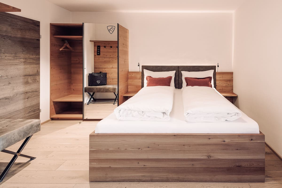 A bedroom with a wooden bed frame, a wardrobe with a coat hanging, and a bench with a bag.