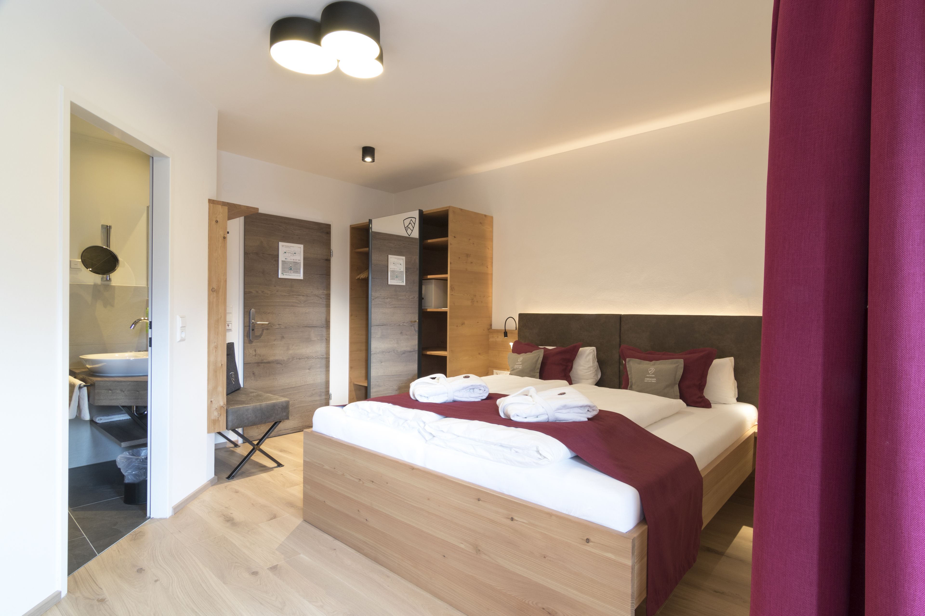 Quarto de hotel moderno com cama de madeira, armário embutido, banheiro com pia e espelho, e luminária no teto.