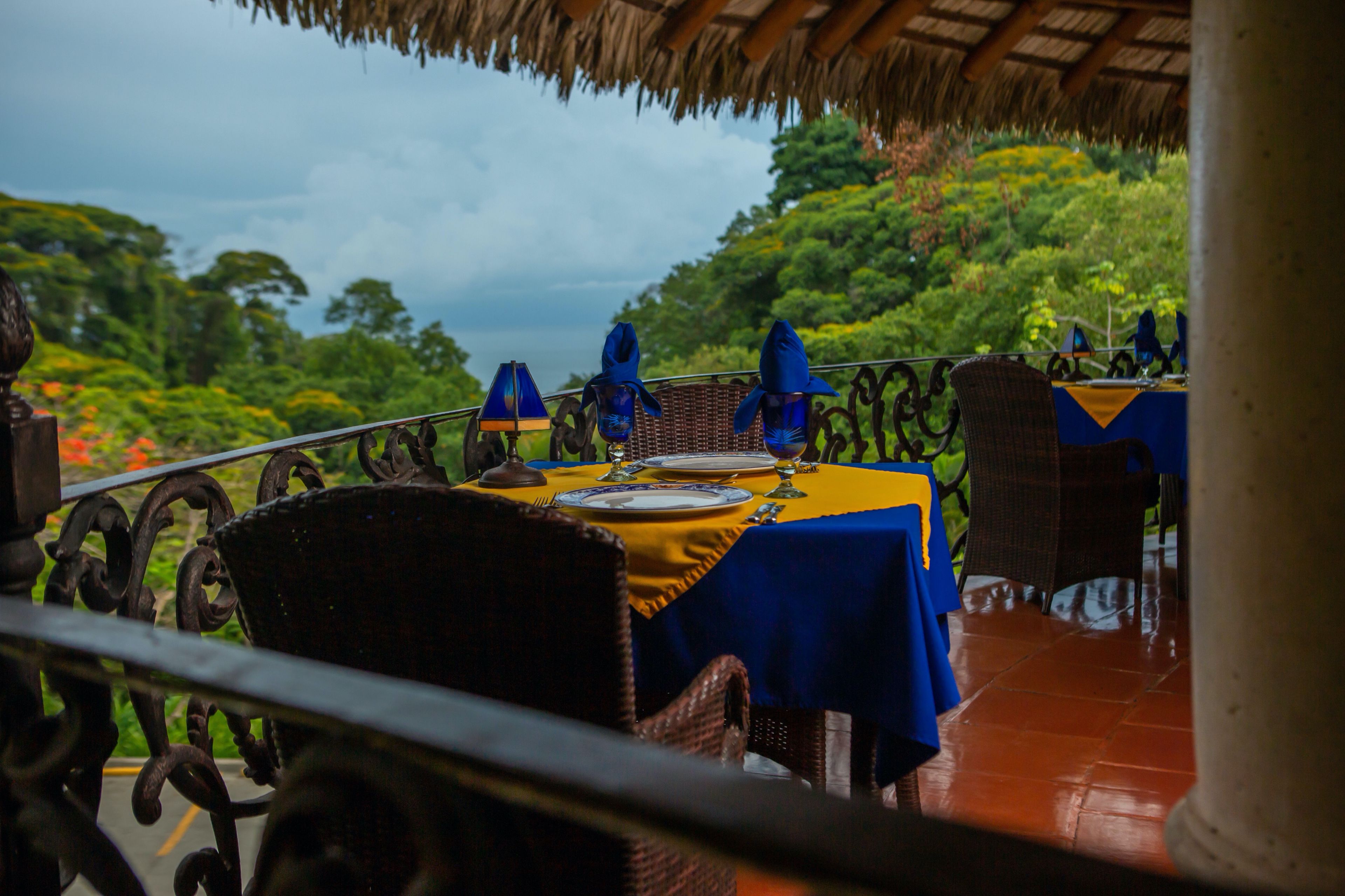 An outdoor dining area with a table set for two, surrounded by wicker chairs and a thatched roof overhead.
