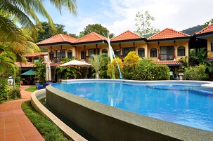 Una piscina con borde de baldosas, rodeada por un edificio con varios balcones y un jardín tropical.