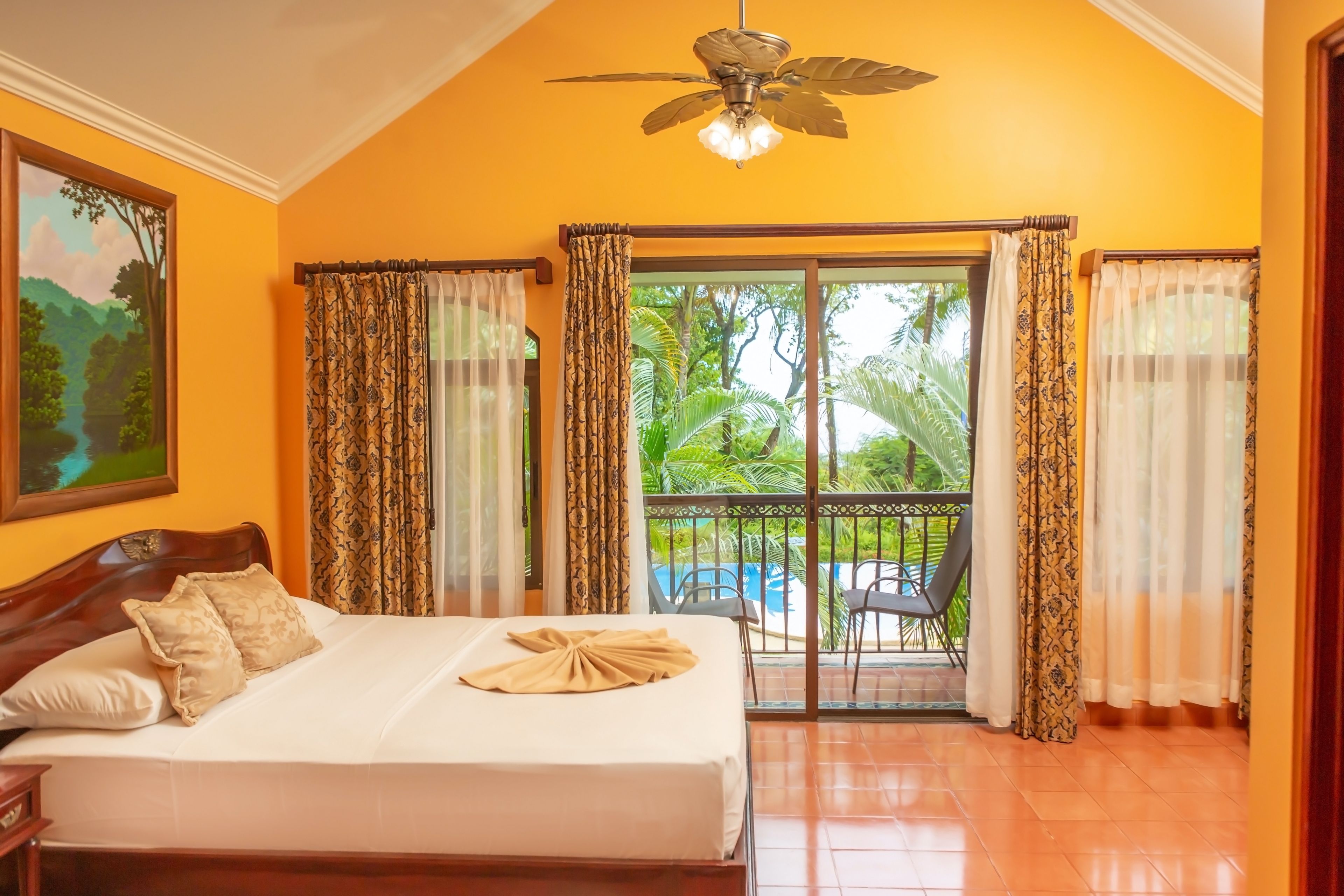 Une chambre d’hôtel avec un lit, un ventilateur de plafond, un tableau et une vue sur une piscine et un espace verdoyant.