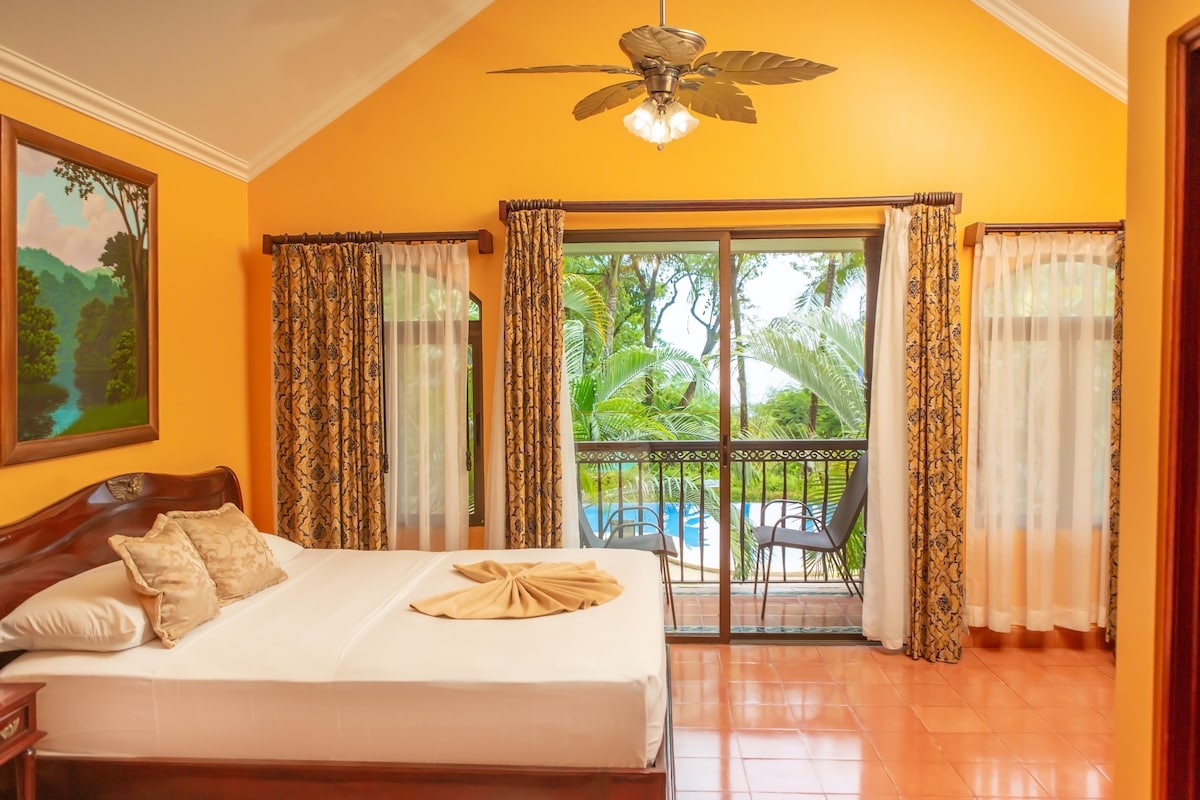 A hotel room with a bed, a ceiling fan, a painting, and a view of a pool and greenery.