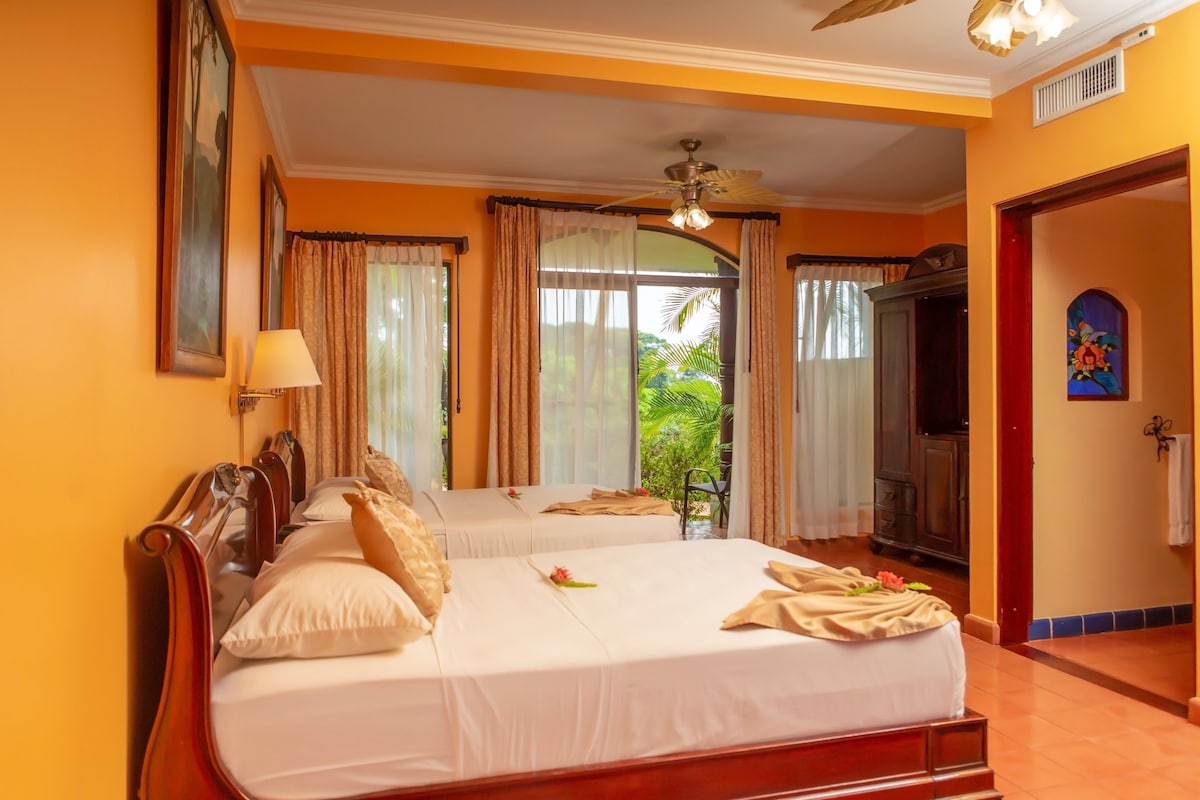 A hotel room with two beds, a ceiling fan, and a view of greenery outside.