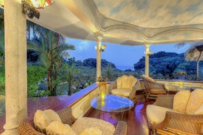 A covered outdoor seating area with wicker furniture and a glass table, overlooking a lush garden and distant hills. - Hotel Cuna Del Angel (Bahía Ballena)