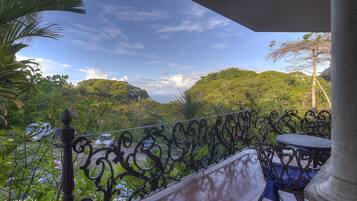 Uma varanda com grade de ferro forjado, uma mesa com toalha azul e vista para uma paisagem verdejante com colinas e céu claro.
