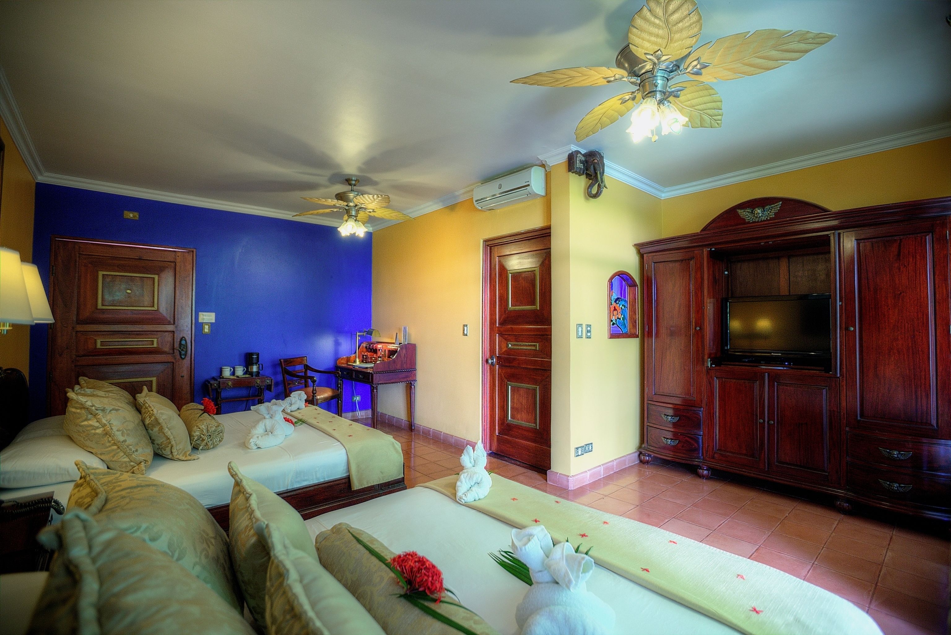 Habitación de hotel con dos camas, televisor, ventilador de techo y un armario de madera.