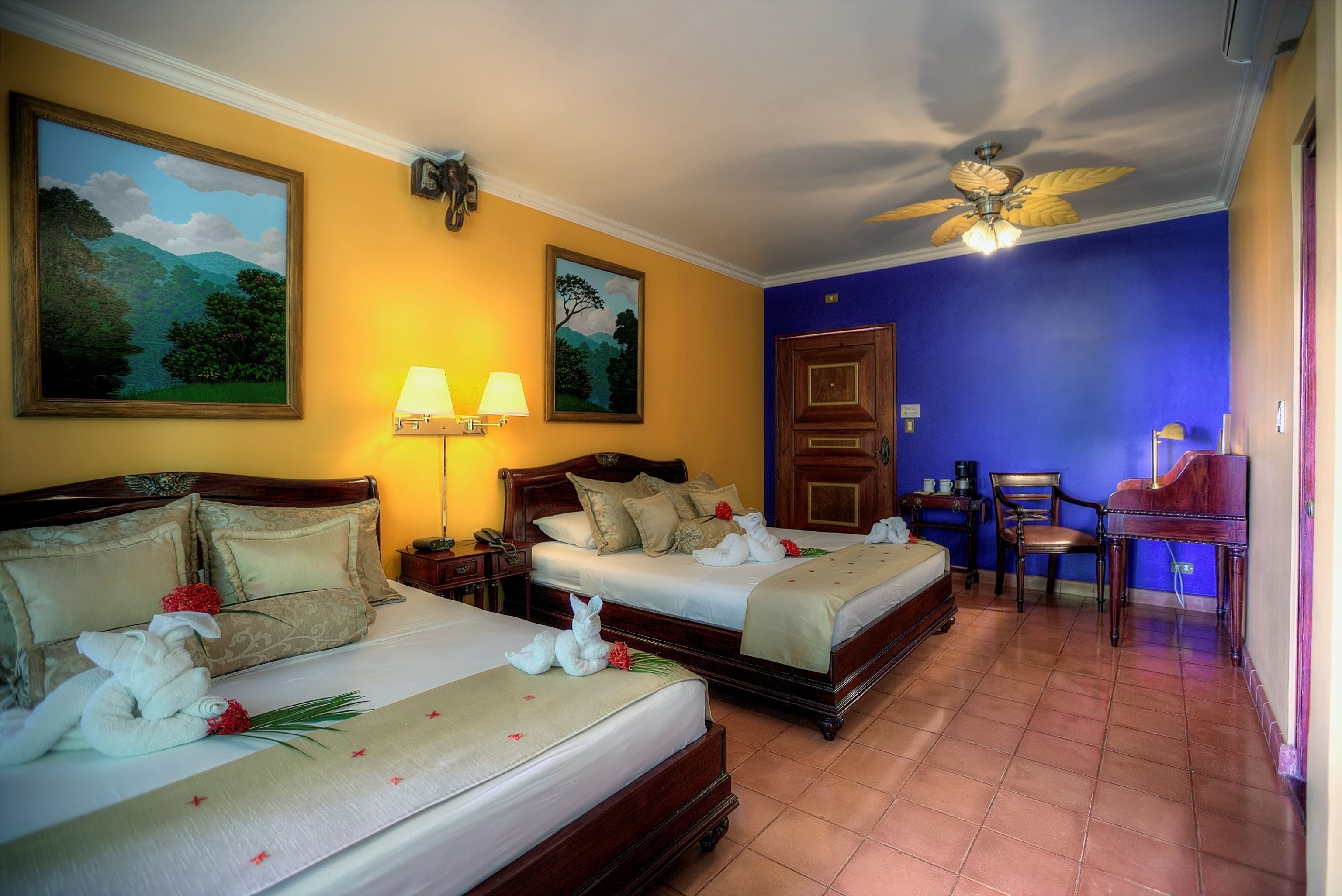 A hotel room with two beds, a desk, a piano, and a ceiling fan.