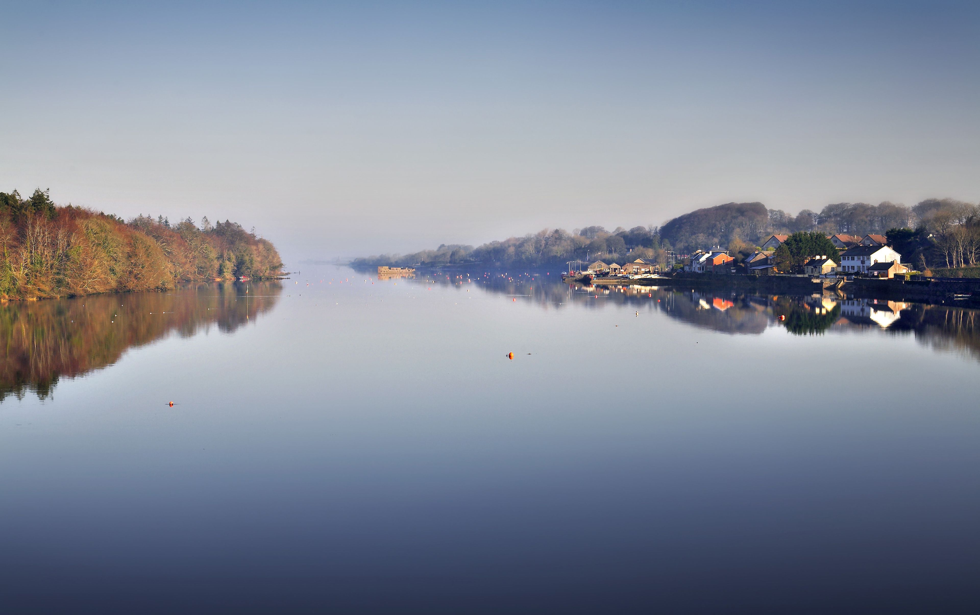 A calm lake with a reflection of houses and trees, surrounded by a forested area.