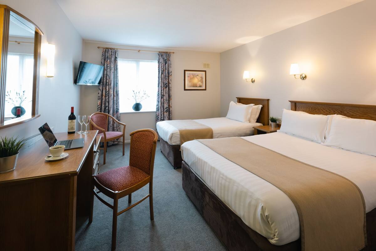 A hotel room with two beds, a desk with a laptop, a chair, a TV, and a window with curtains.