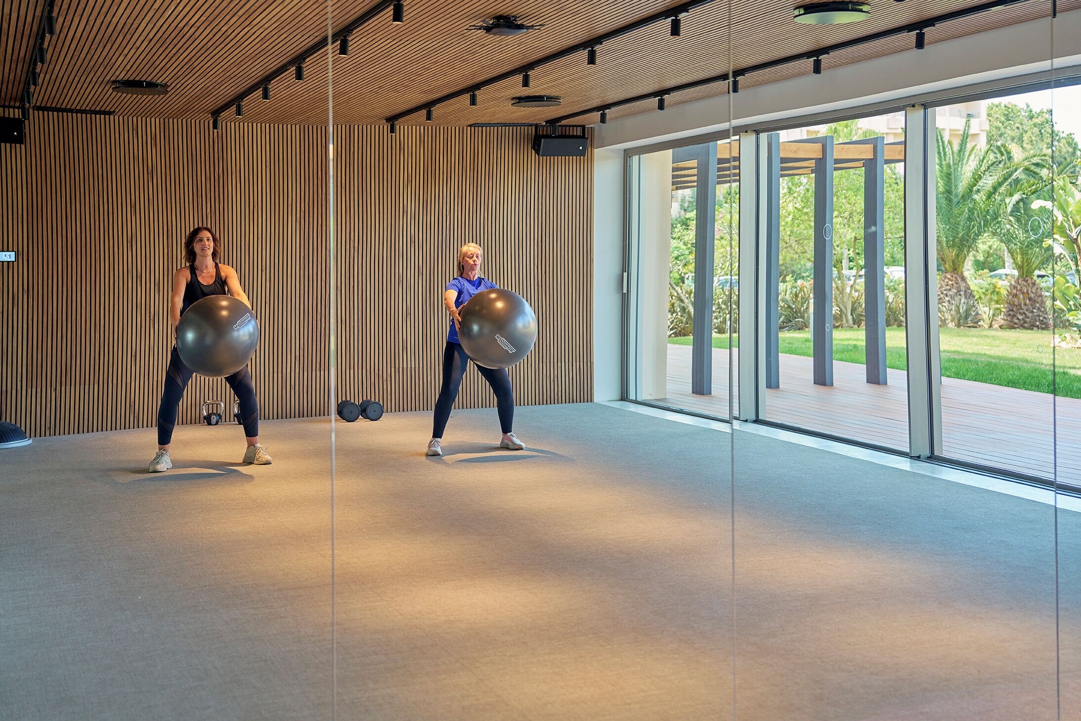 A modern gym with wooden panel walls and large windows offering a view of greenery outside.