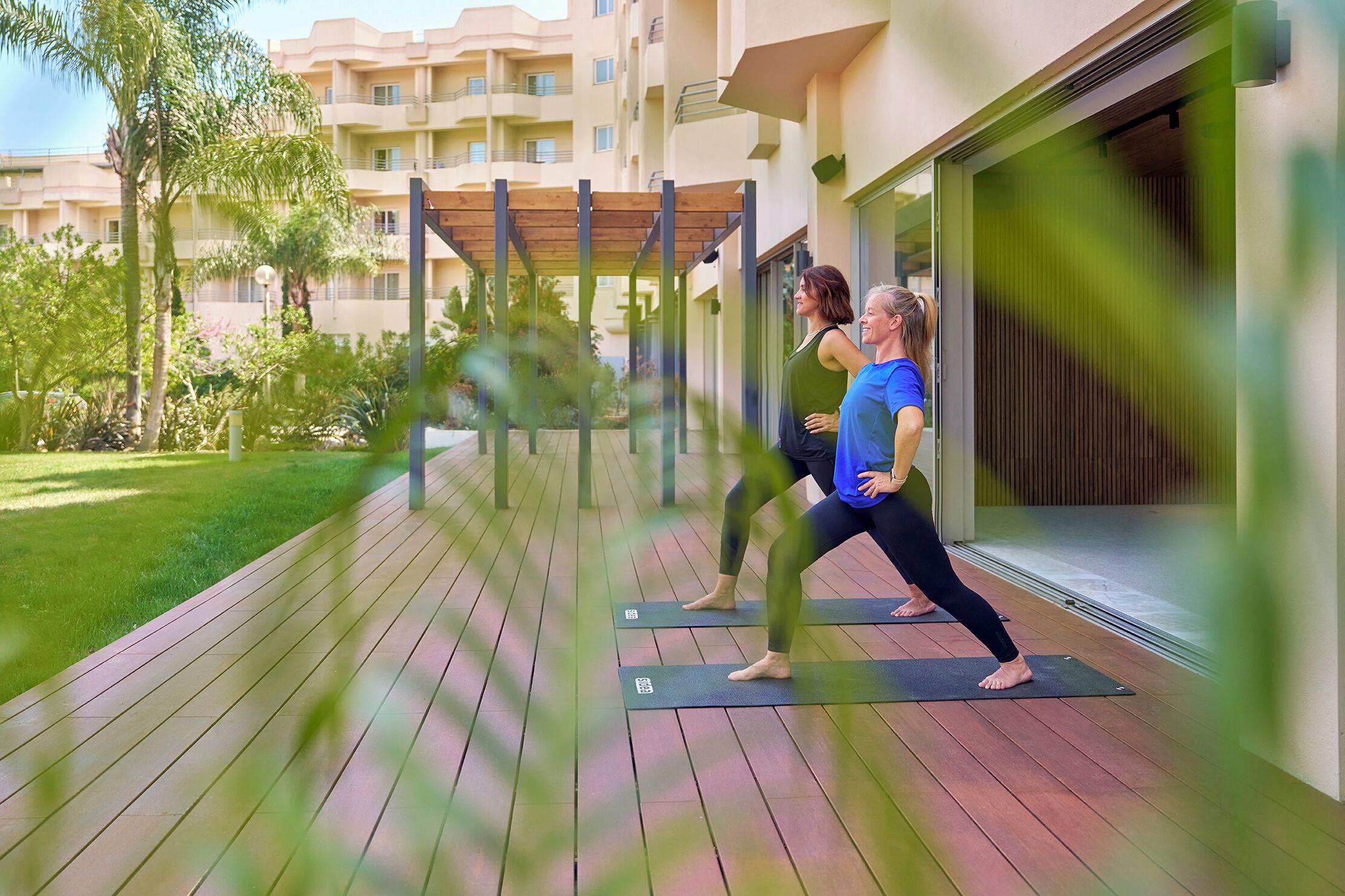 Dos personas practicando yoga en terrazas de madera al aire libre, con un edificio al fondo.
