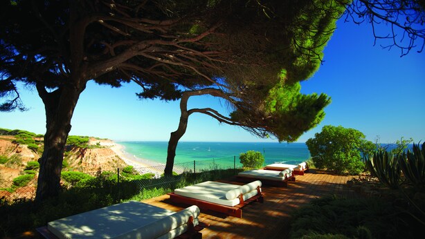 A secluded outdoor lounge area with wooden flooring and multiple beds arranged in a row, overlooking a beach and the sea.