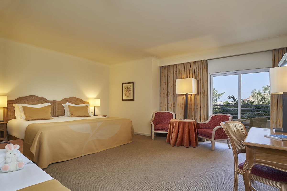 Een hotelkamer met een groot bed, twee nachtkastjes, een bureau, een stoel en uitzicht op het buitenleven door een groot raam.