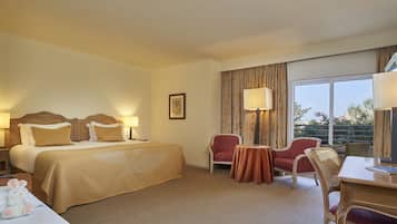 Habitación de hotel con una cama grande, dos mesitas de noche con lámparas, un escritorio con silla, una pequeña mesa con silla y un ventanal con cortinas.