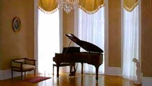 A grand piano in a spacious room with a chandelier, large windows with curtains, and a framed picture on the wall. - The Confederate House (Savannah)