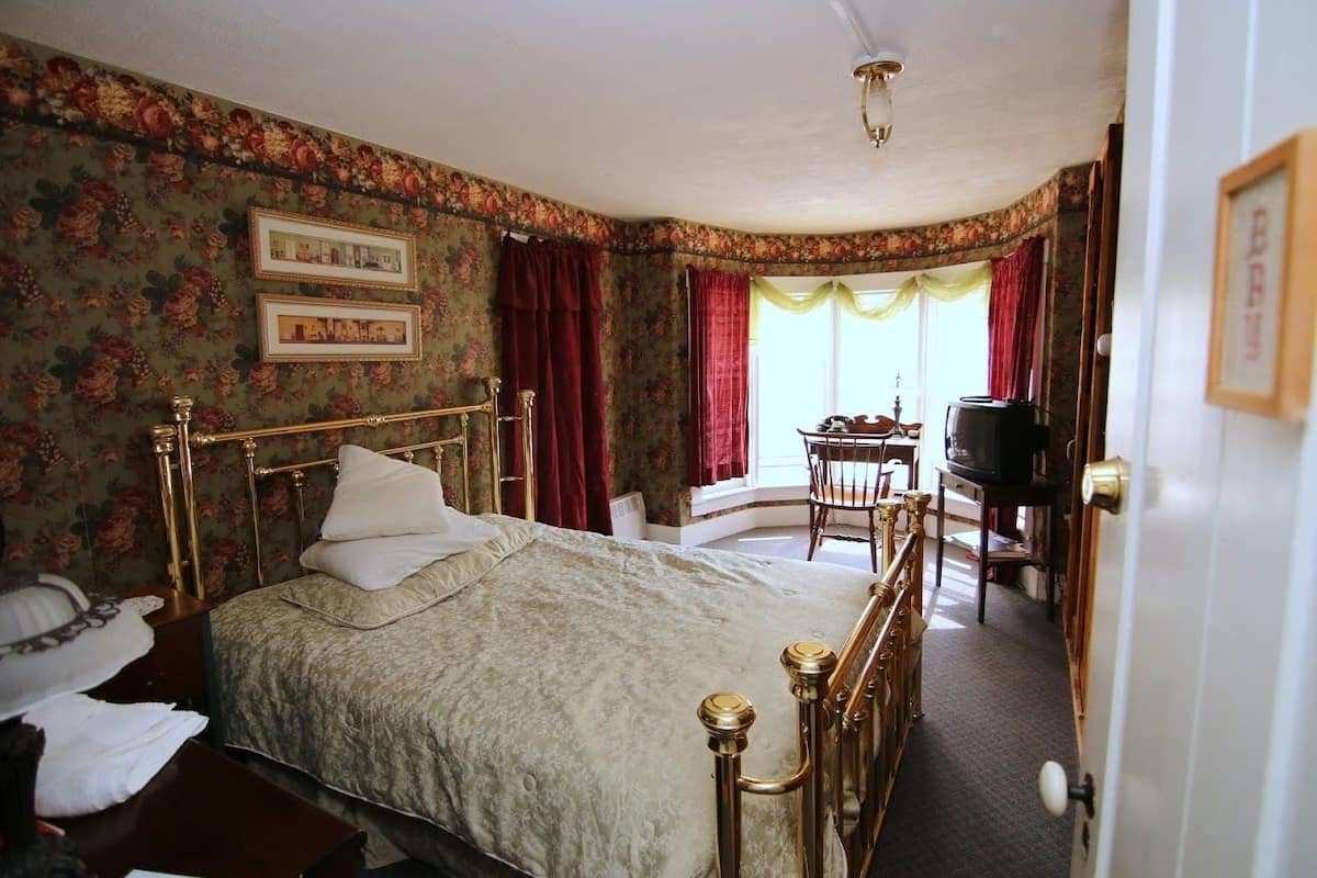 A bedroom with a four-poster bed, floral wallpaper, a desk with a chair, and a television.