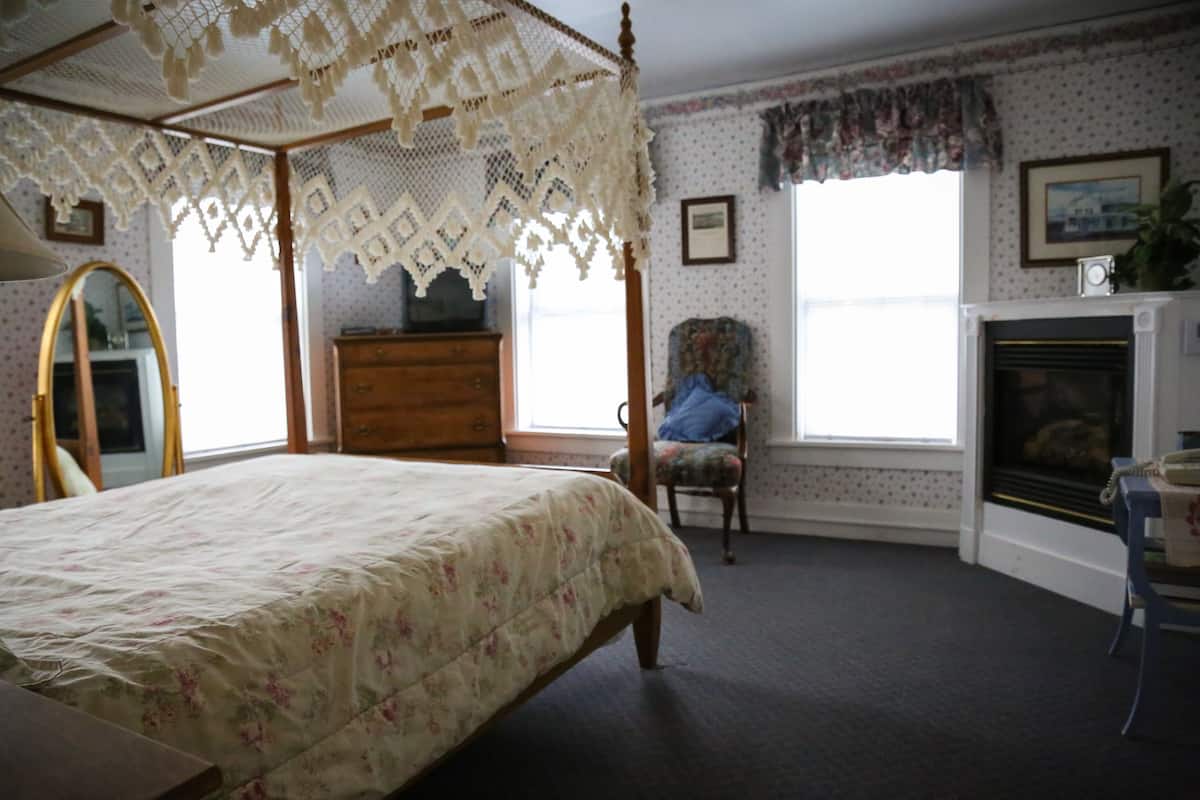 A bedroom with a four-poster bed, a dresser, a chair, a fireplace, and a window with curtains.