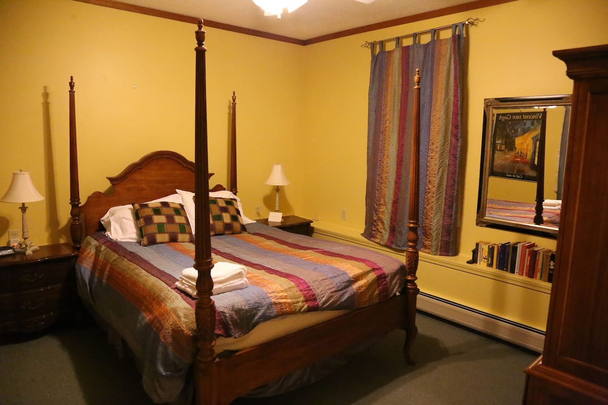 A four-poster bed with a colorful quilt, two bedside tables with lamps, a large mirror, and a bookshelf with books.