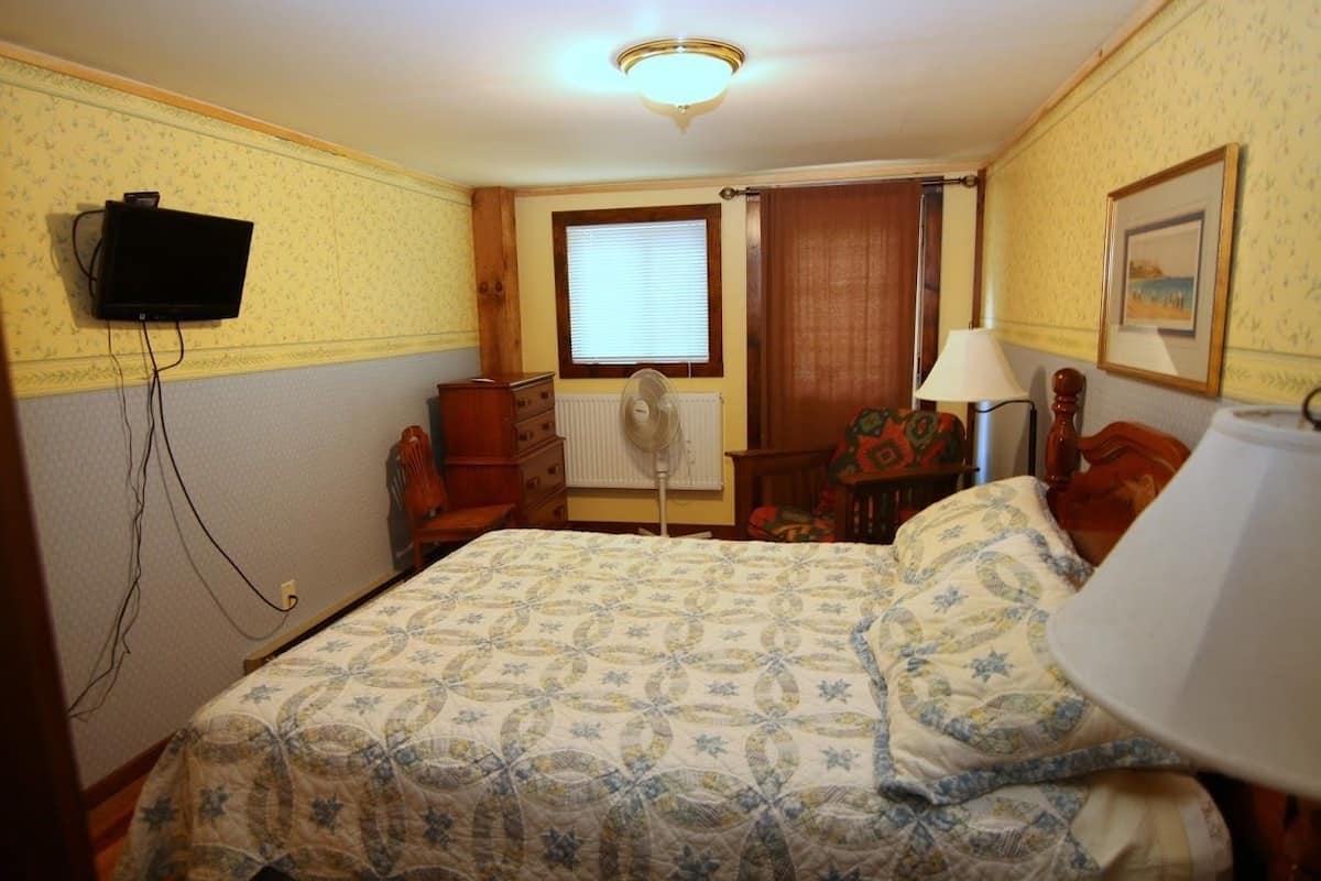 A bedroom with a bed, a television mounted on the wall, a window with blinds, a radiator, a lamp, and a framed picture on the wall.