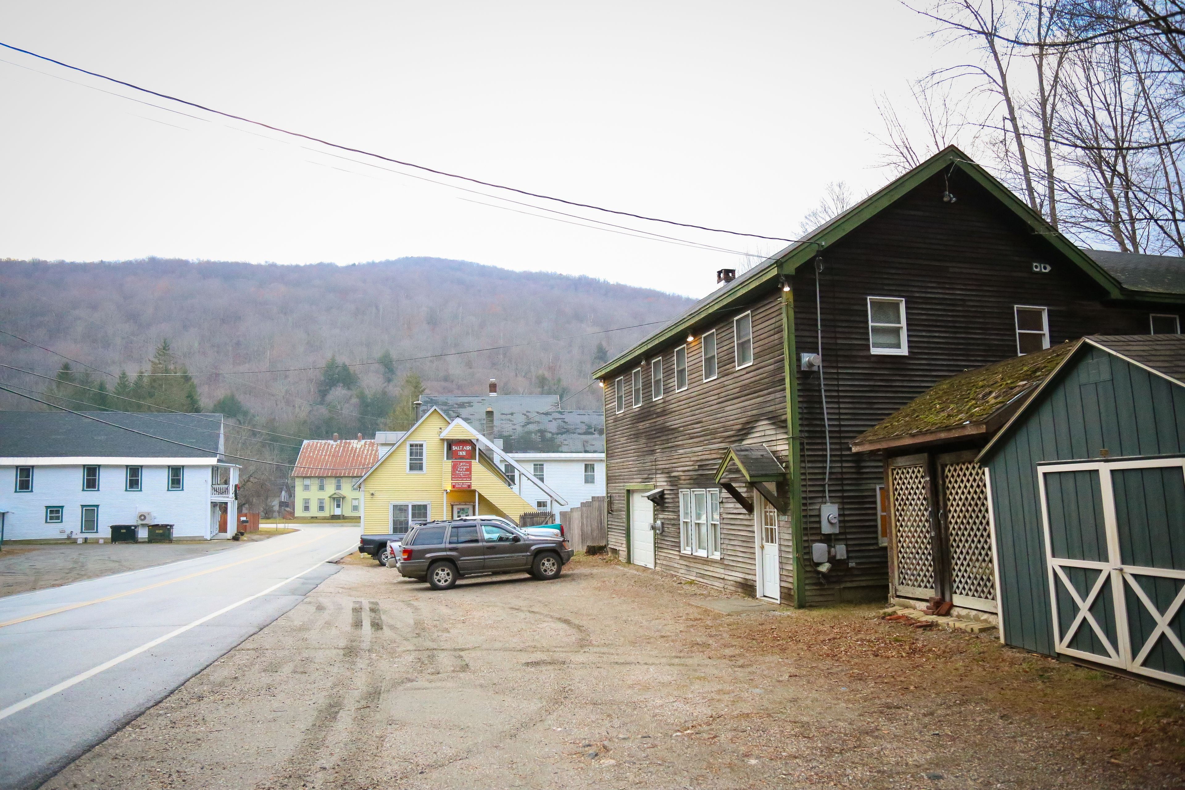 A rural road with a parked SUV, a wooden building with a green door, and a yellow building with a red sign.