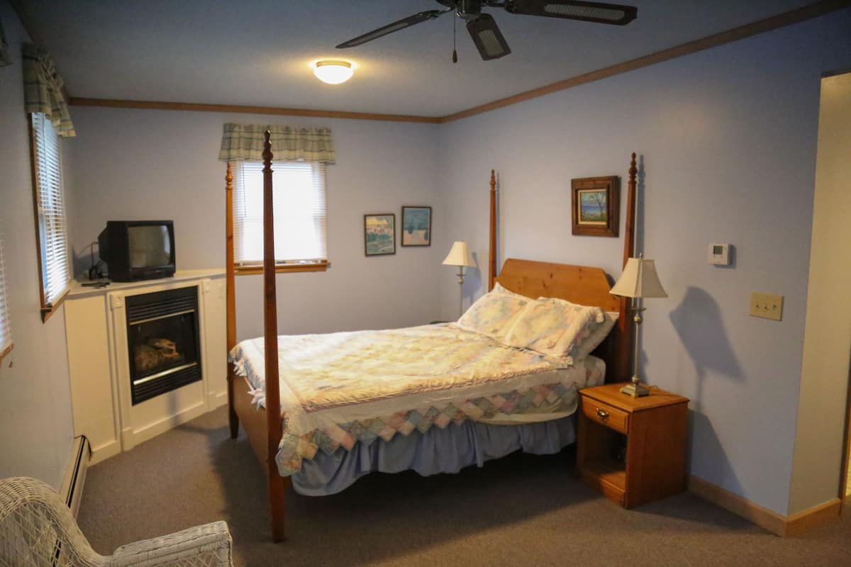 A bedroom with a four-poster bed, a nightstand with a lamp, a television on a stand, and a fireplace.
