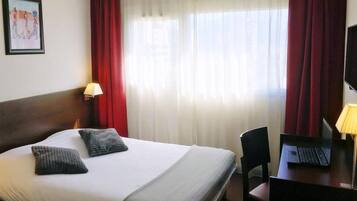 Habitación de hotel con cama, escritorio, laptop, silla, lámpara y televisor.