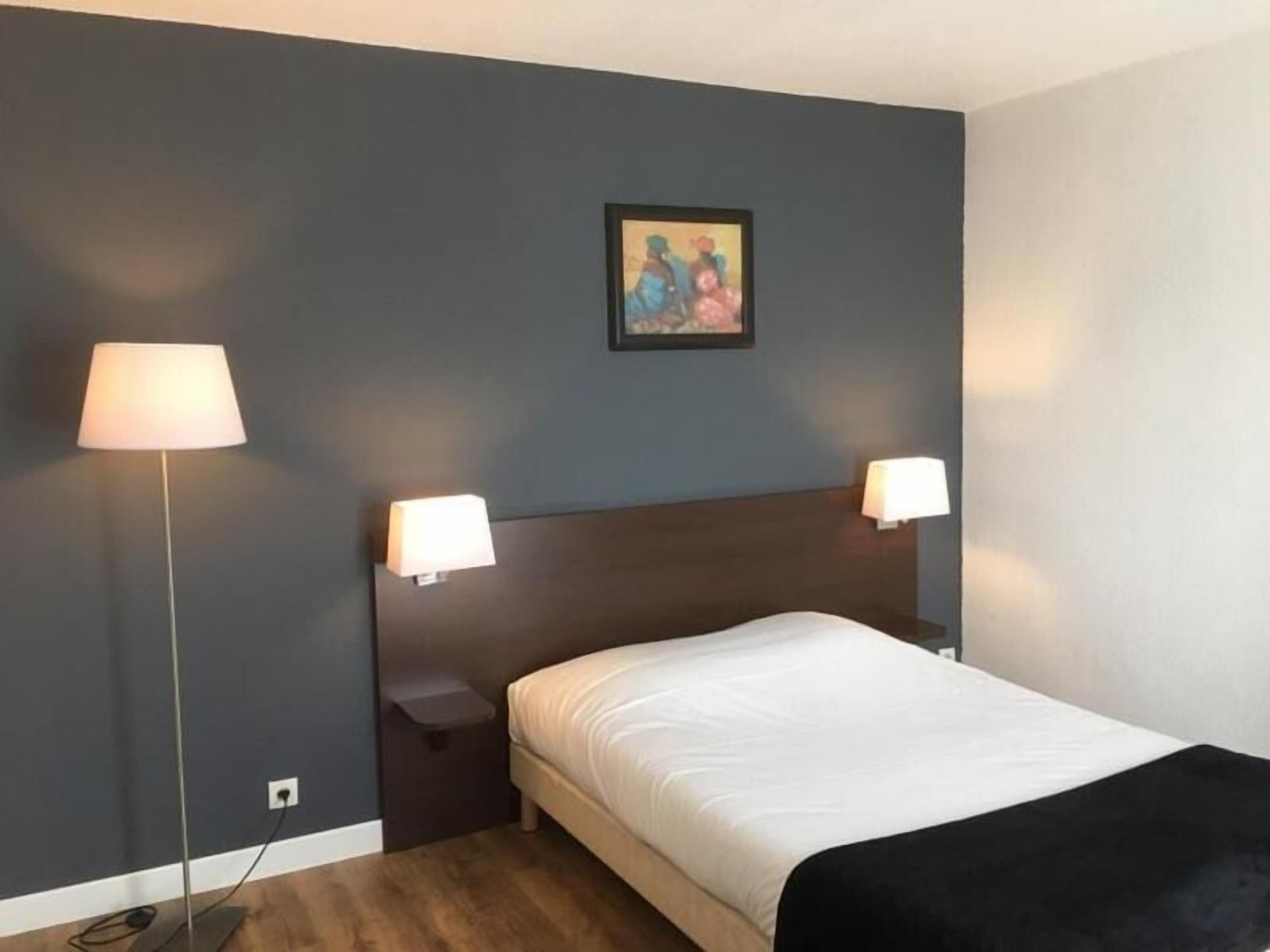 Une chambre d’hôtel avec un mur bleu foncé, un lit avec une literie blanche, deux lampes de chevet et un petit tableau au-dessus du lit.