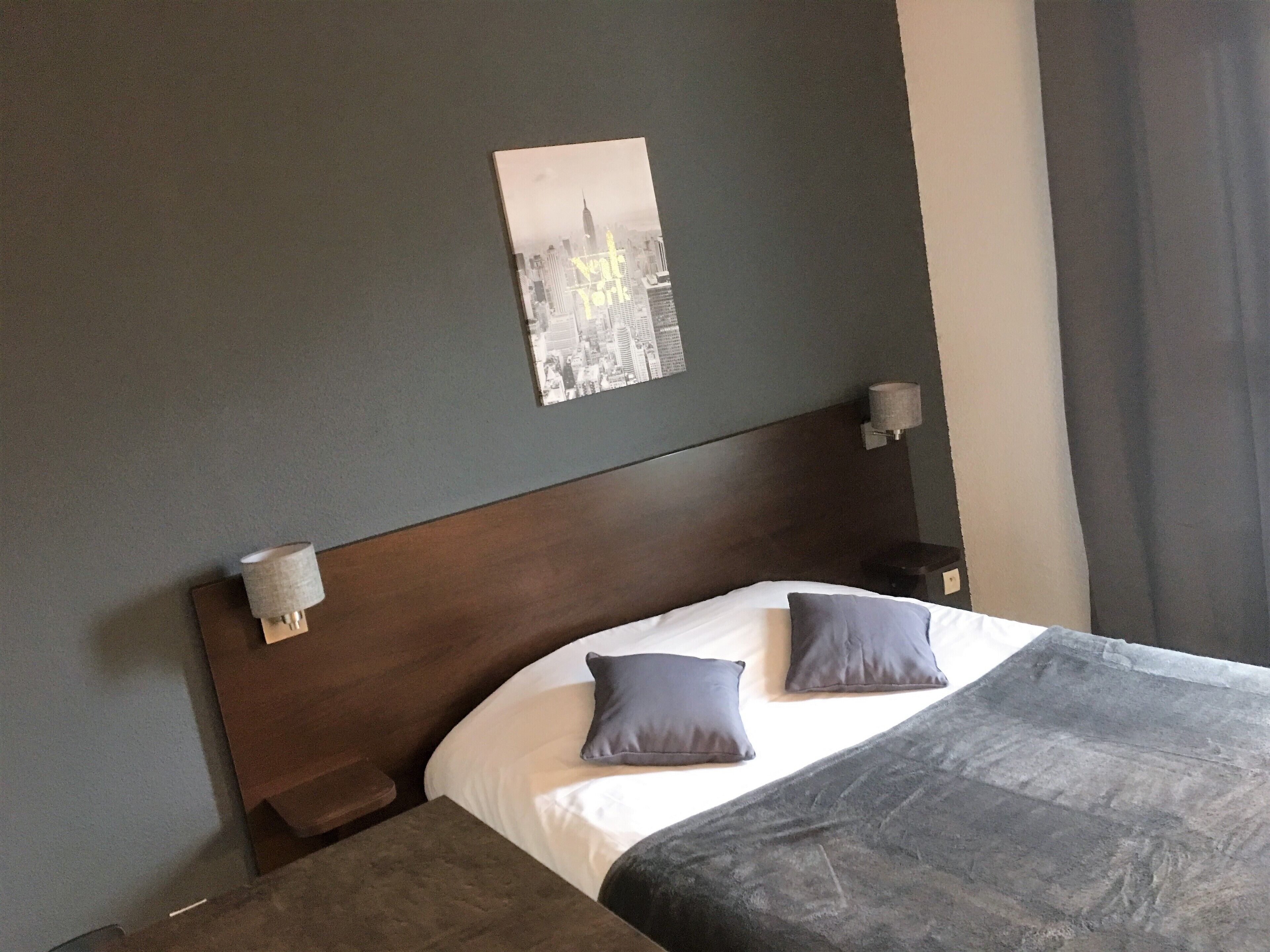 Quarto de hotel com uma cama grande, duas almofadas, uma mesa de cabeceira, uma luminária na parede e um quadro enquadrado.