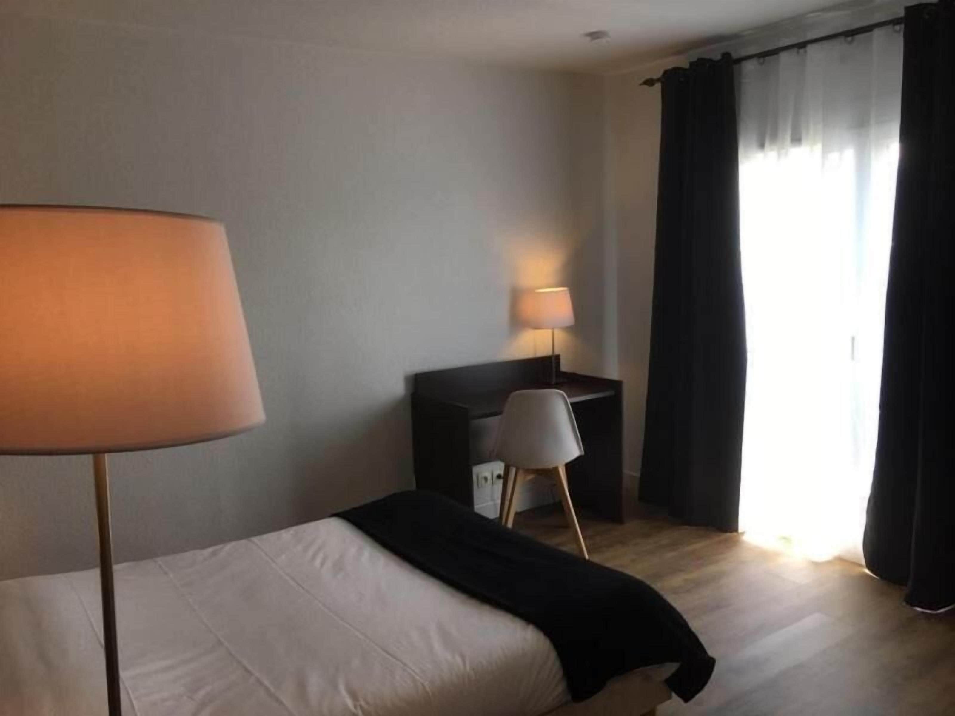 Una habitación de hotel con una cama, un escritorio, dos lámparas y un ventanal con cortinas.