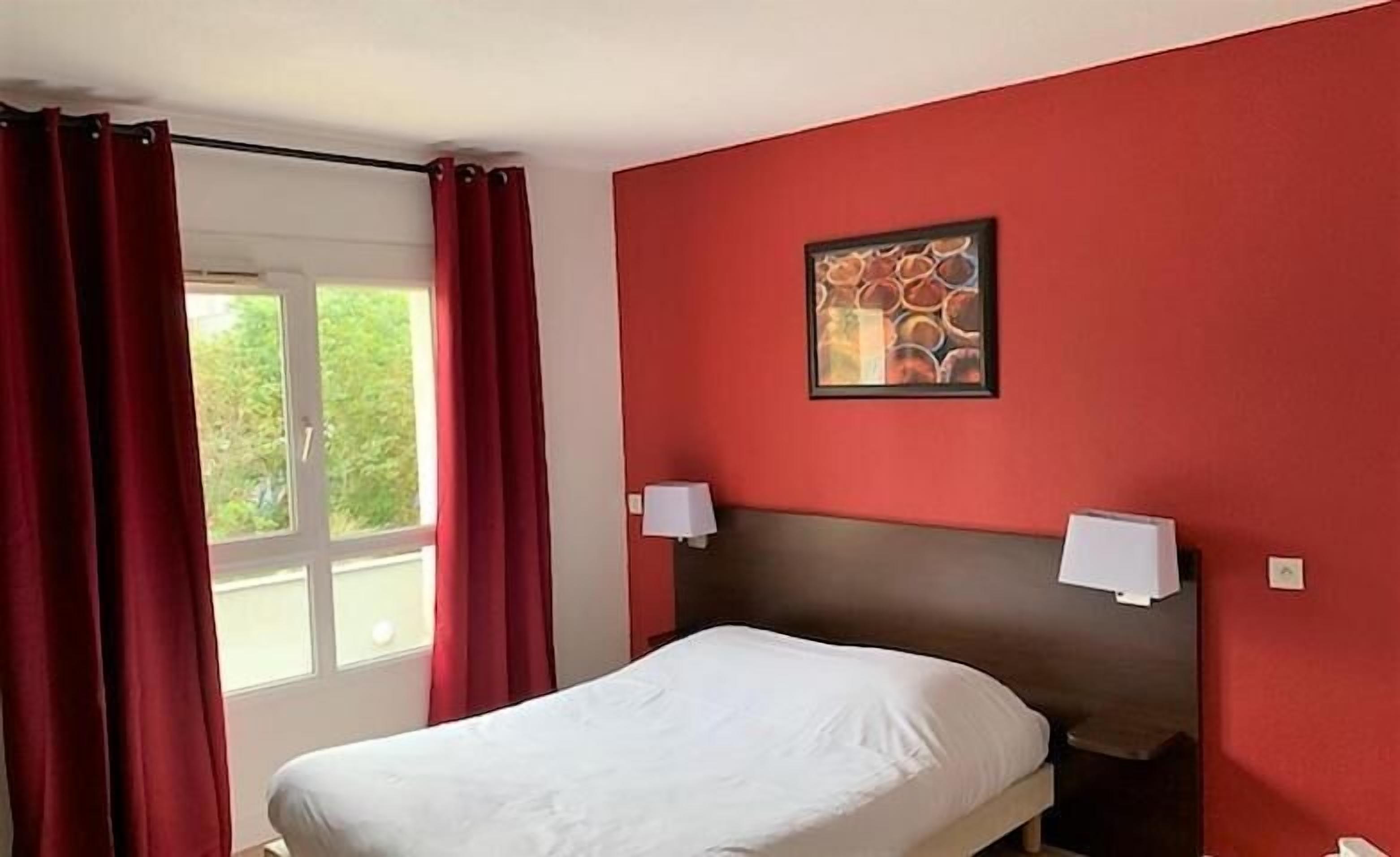 Una camera d'albergo con un letto, pareti rosse, una finestra con tende e un quadro appeso al muro.