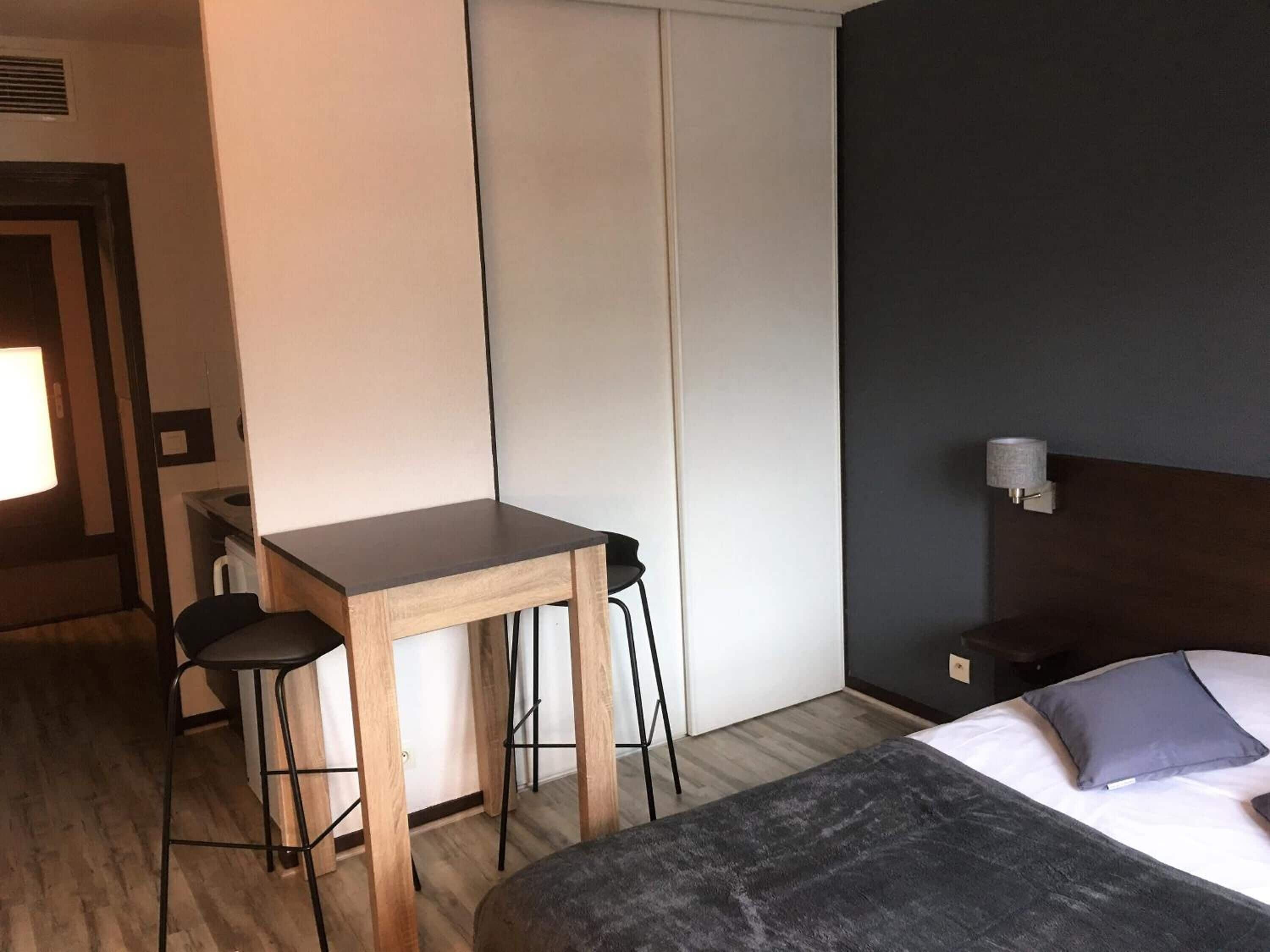 A hotel room with a bed, a desk with two chairs, a nightstand, and a wall-mounted lamp.