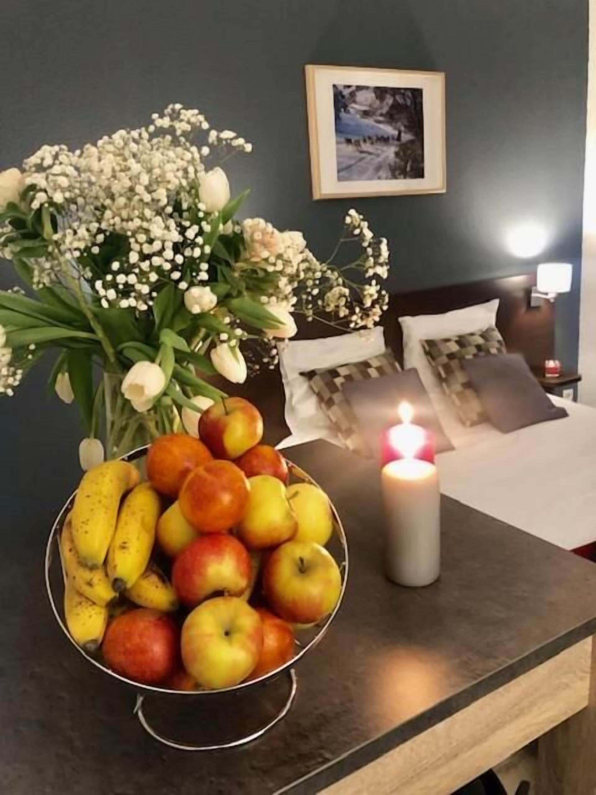Een netjes gerangschikte fruitschaal met appels en bananen, een brandende kaars en een ingelijste foto op een nachtkastje in een hotelkamer.