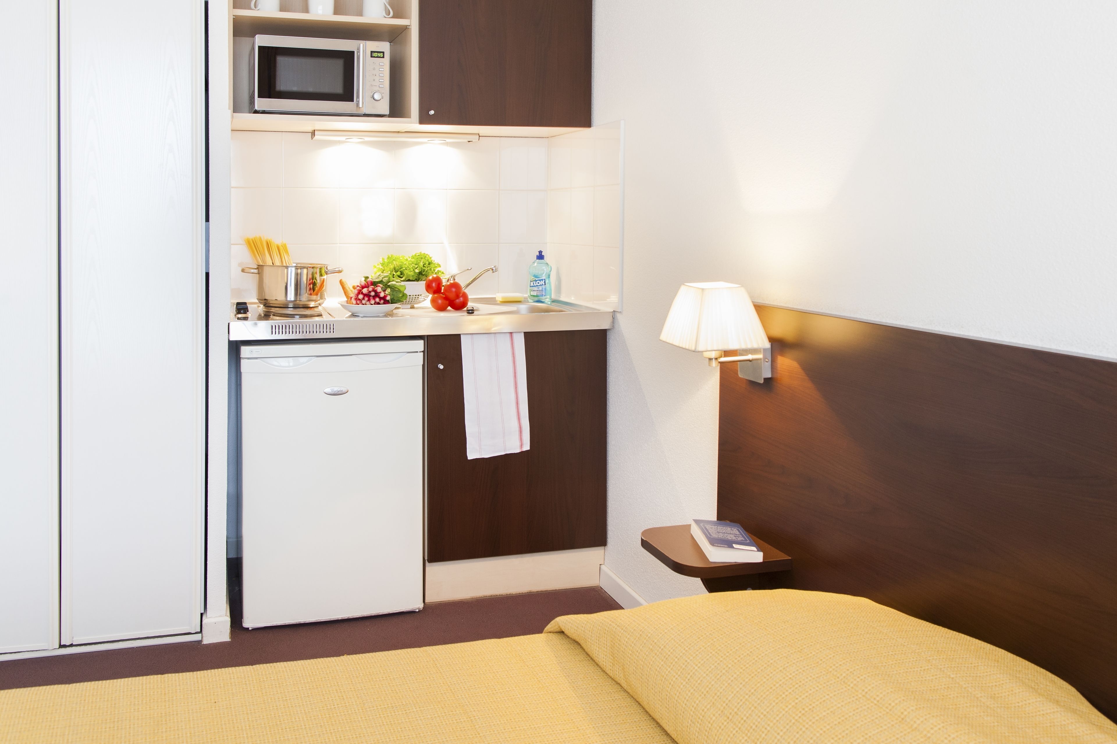 A compact hotel room with a kitchenette, including a microwave, a small fridge, and a sink. There is a bedside table with a lamp and a book.