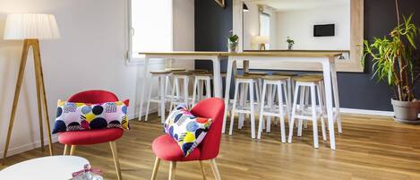 A modern living room with a dining area, featuring a wooden floor, a red chair with a patterned cushion, a small round table, and a bottle of Bonbons.