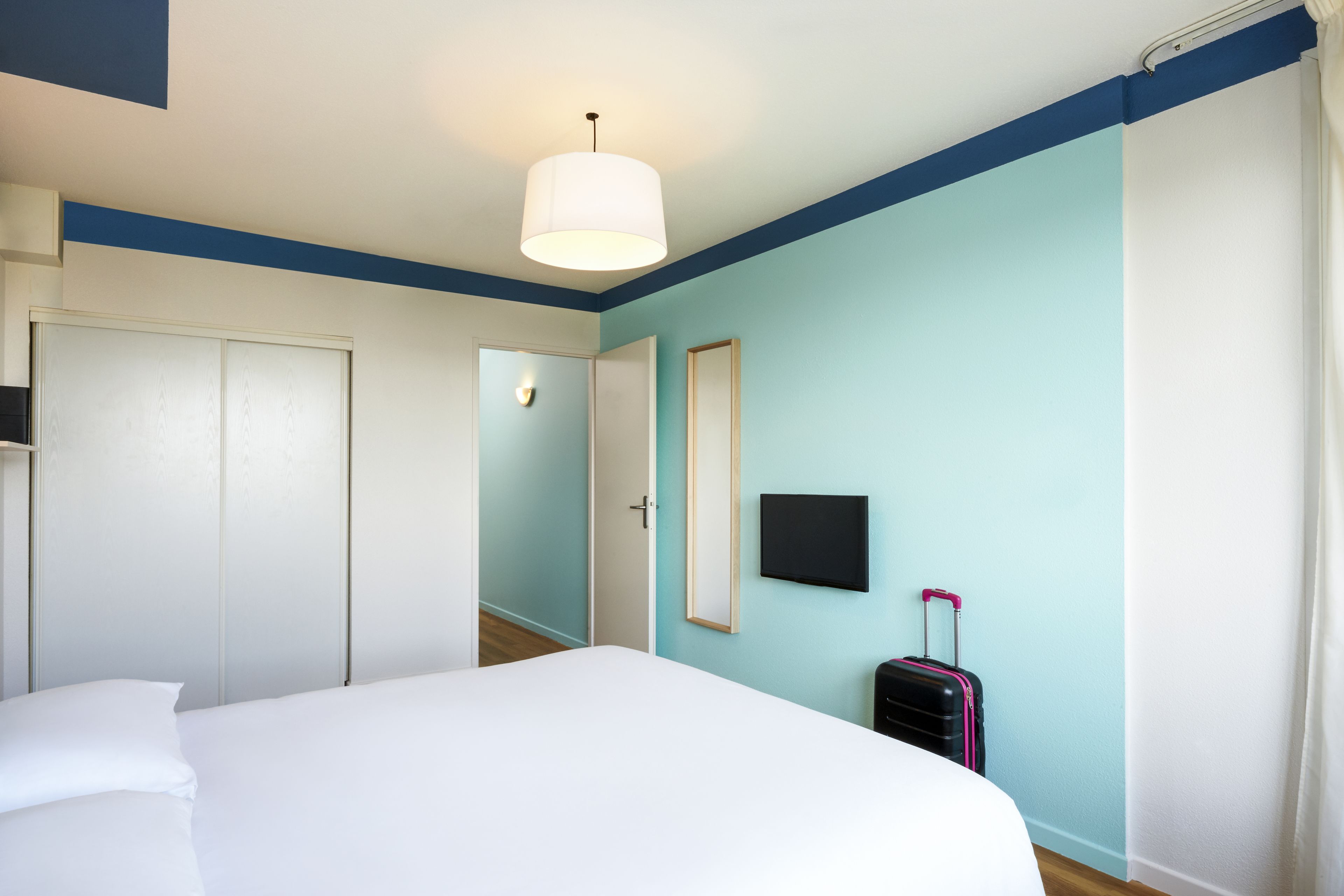 A hotel room with a bed, a suitcase, a flat-screen TV, and a door.