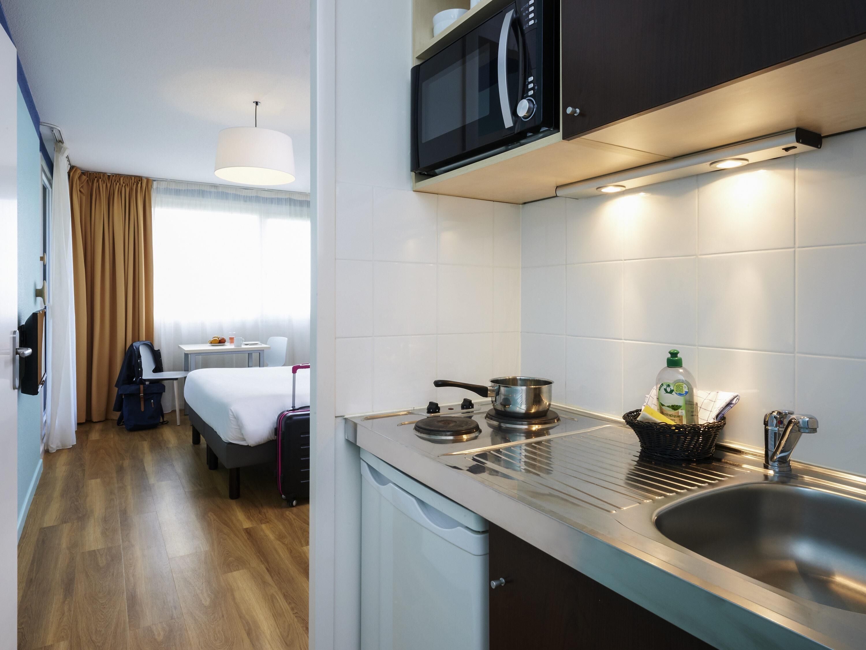 A compact hotel room with a kitchenette, a bed, a desk, and a window with curtains.