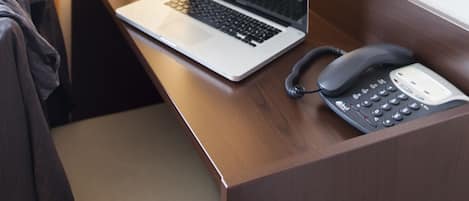 A hotel room with a wooden desk, a laptop, a telephone, a vase with flowers, and a framed picture on the wall.