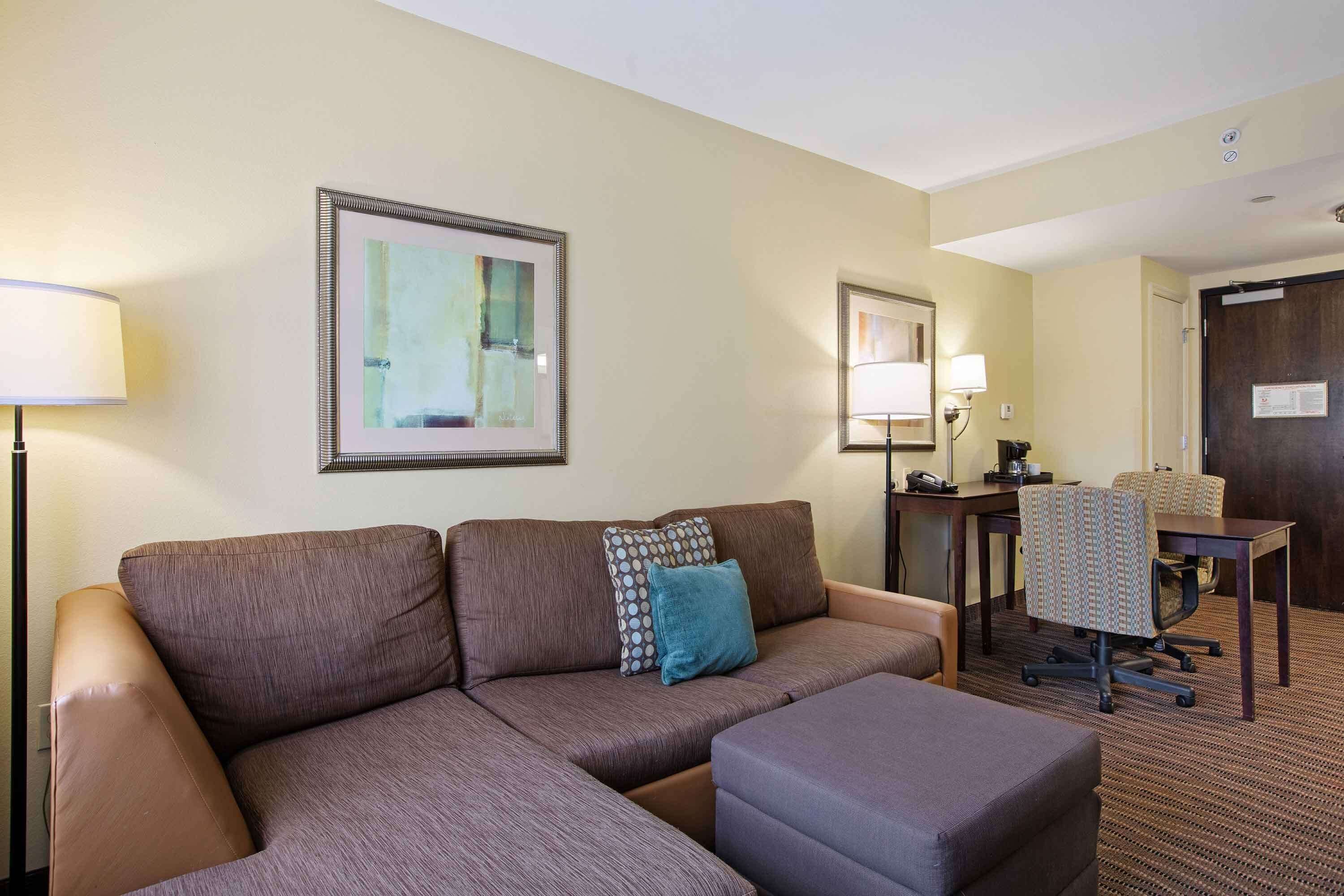 A hotel room with a sectional sofa, a small table, two chairs, and a desk with a lamp.