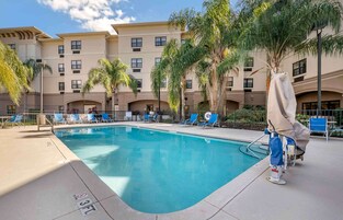 Una piscina con sillones de descanso y palmeras, rodeada por un edificio de varios pisos con balcones.